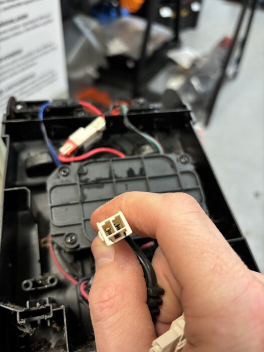 Hand håller ett vitt AMP-kontaktdon med två stift, bakgrund av kablar och en svart plastkomponent.