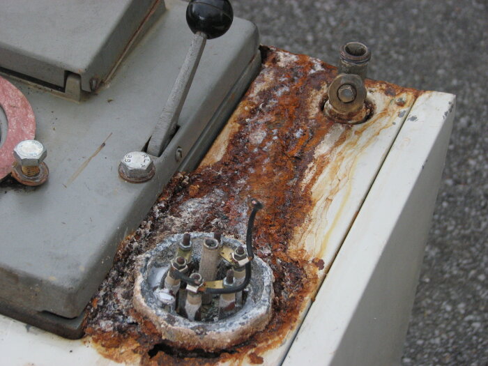 Rostig del av en maskin med vents och spakar på ytan, omgiven av tydliga tecken på korrosion och slitage.