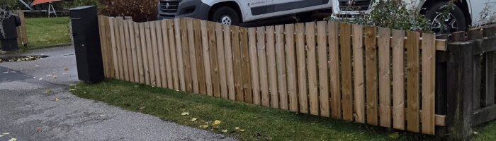 Trästaket med vertikala plankor och stolpar, omgiven av gräs och asfaltväg. En transportbil syns i bakgrunden.