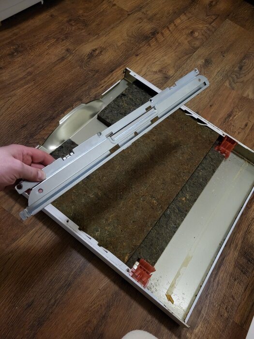 Metallskena och diskmaskinens frontkåpa, demonterad och liggande på trägolv, med problem att återmontera metallskenan korrekt.