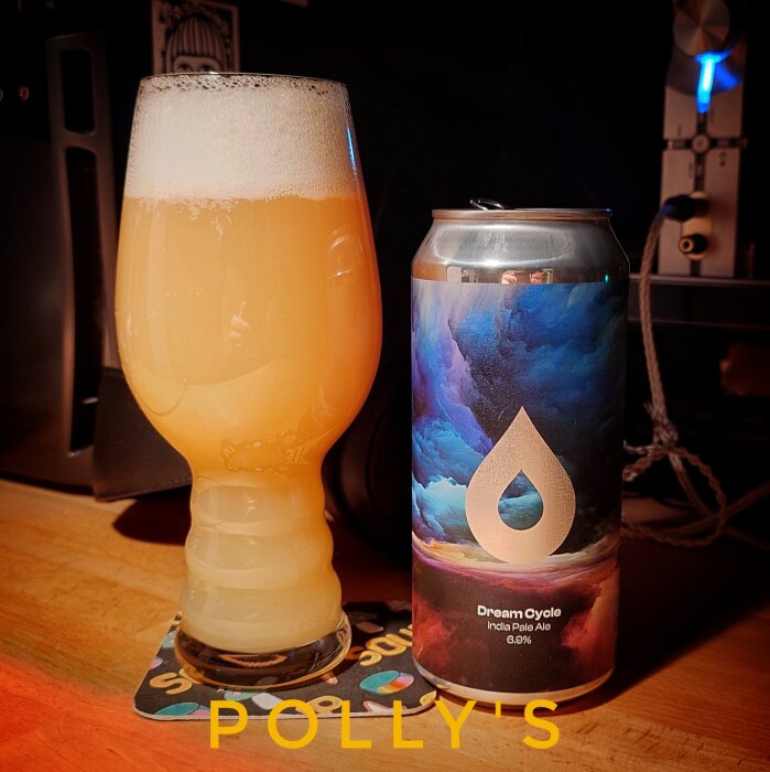 Ölglas med skummande IPA och en burk Polly's Dream Cycle IPA på bord med underlägg.
