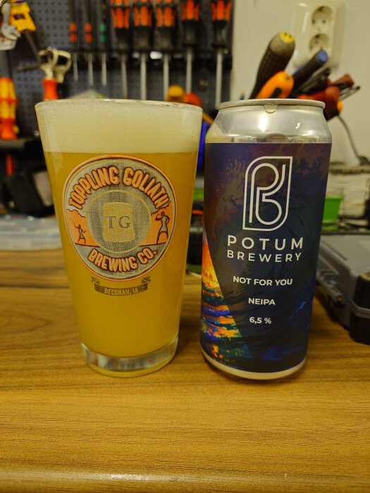Glas med ljus öl från Toppling Goliath och burk med Potum Brewery NEIPA, 6,5 %, på ett arbetsbord framför verktyg.