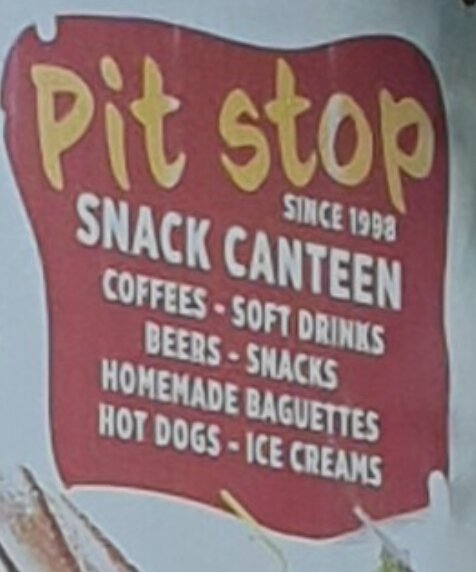 Skylt med text: "Pit stop snack canteen since 1998". Erbjuder kaffe, läsk, öl, snacks, baguetter, korv med bröd och glass.
