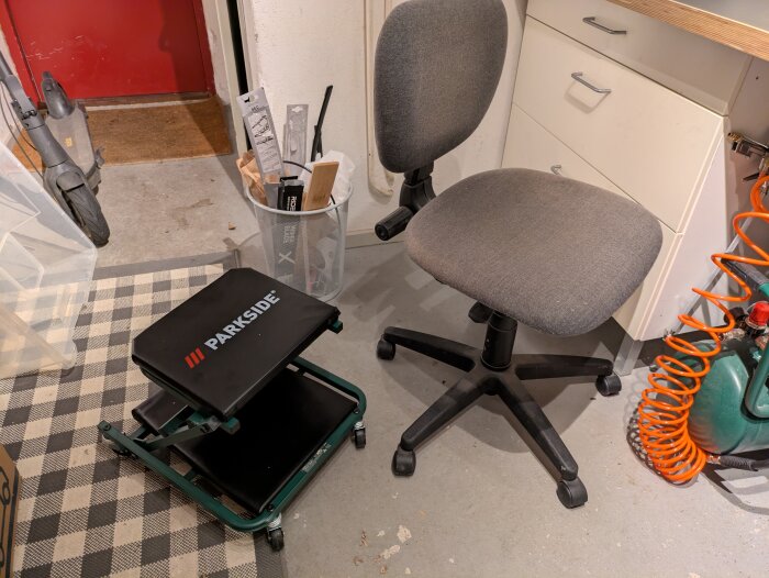 Rullande verkstadsstol märkt "Parkside" bredvid en grå kontorsstol, stående på betonggolv med orange tryckluftsslang i bakgrunden.