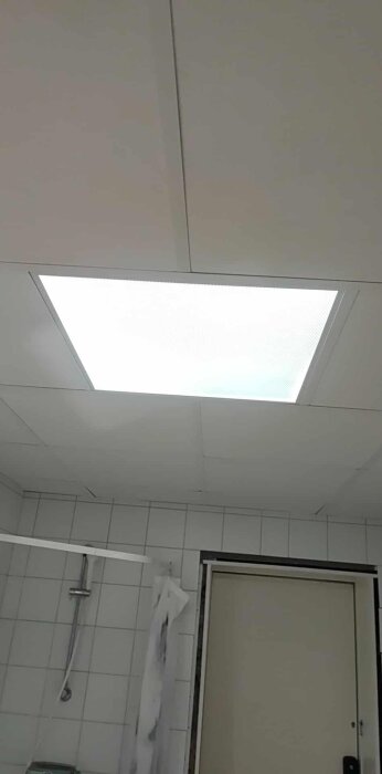 LED-lampa i taket lyser, del av badrum syns med dusch och kakelväggar.