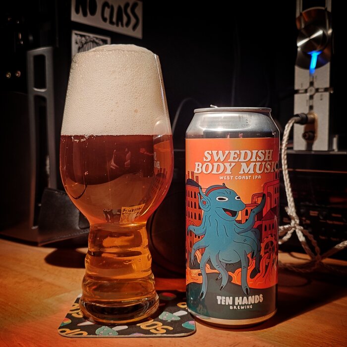 Ett glas med skummande öl bredvid en burk med etiketten "Swedish Body Music" West Coast IPA, på ett bord.