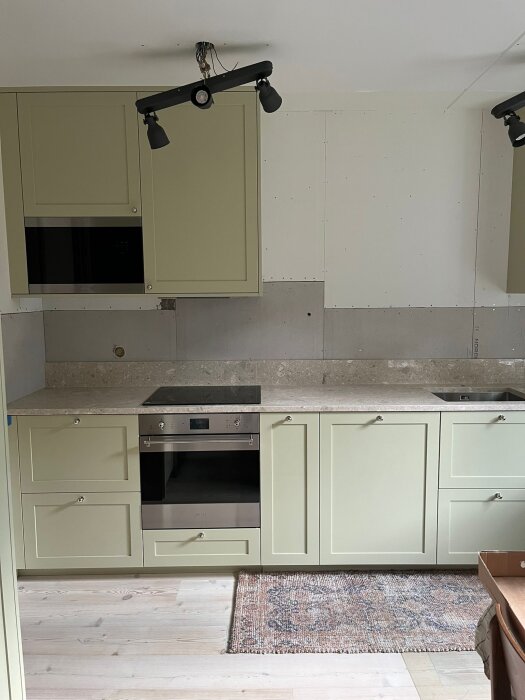 Kök med gröna skåp, ovansida i sten och gipsskivor på väggen, där skarvarna mellan skivorna syns. Taklampor utan glödlampor är installerade.