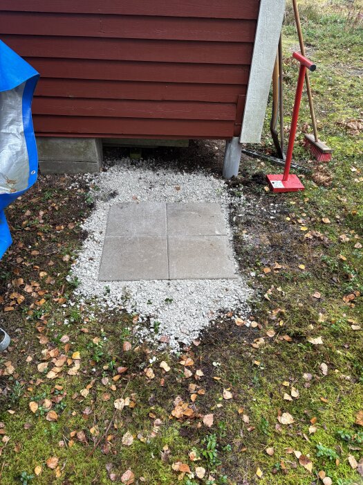 Fundament med plattor och grus vid röd stuga, bas för värmepump. Liggande löv och grönska runt om.