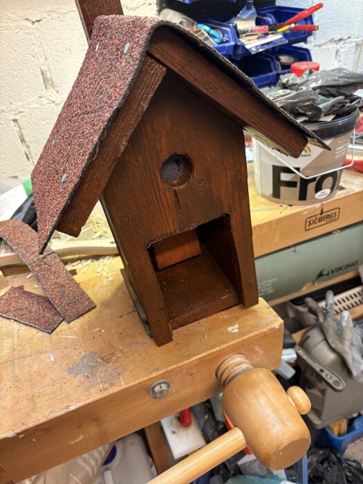 Renoverad fågelholk med nytt takmaterial, placerad på en arbetsbänk med verktyg och material i bakgrunden.