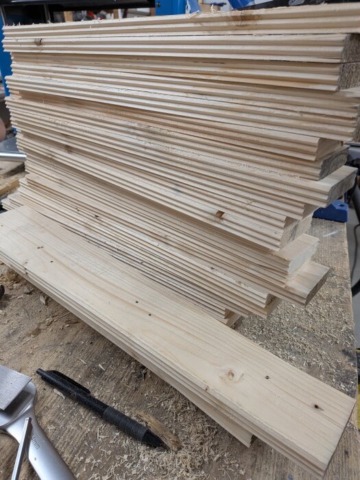 Staplade träbitar av tunt virke på ett arbetsbord, omgivna av träspån och verktyg i en verkstadsmiljö.