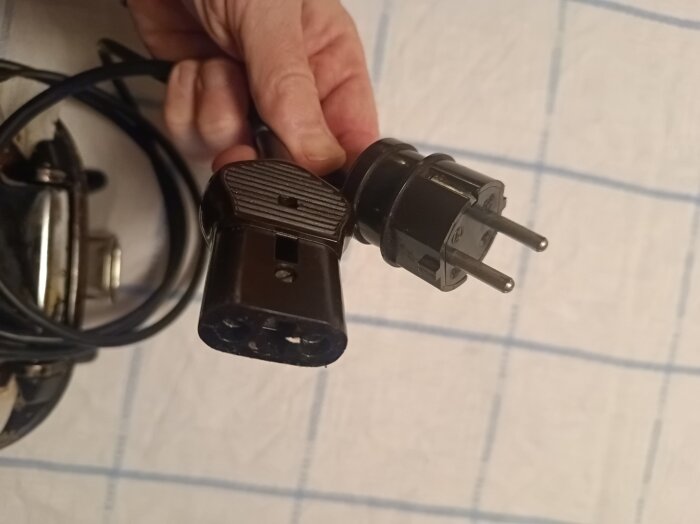 Hand som håller en ojordad kontakt och sladdsticka, på en vit-rutig bakgrund. Frågan gäller om kontakten bör vara jordad.