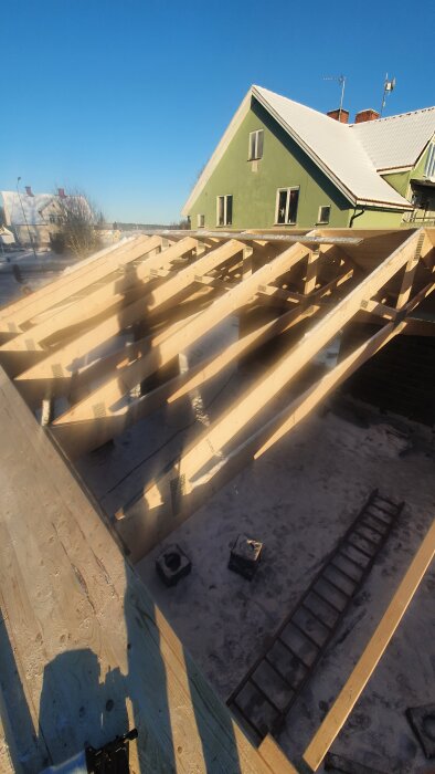 Garage under konstruktion med synliga takbjälkar och en skugga av en person. Grön byggnad i bakgrunden under en klarblå himmel.