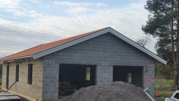 Garage under uppbyggnad med nytt tak och väggar av betongblock, omgivna av träd. Grushög och byggmaterial syns framför garaget.