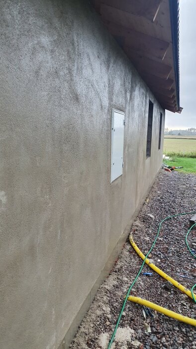 Nyputsad garagevägg med synligt elskåp och fönster, omgiven av grus och slangar; taket syns ovanför.