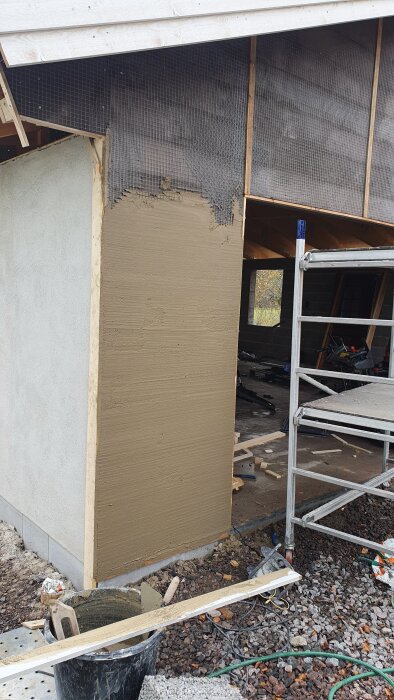 Garagets vägg under putsning med bruk och putsnät, ställning intill och arbetsmaterial på marken.