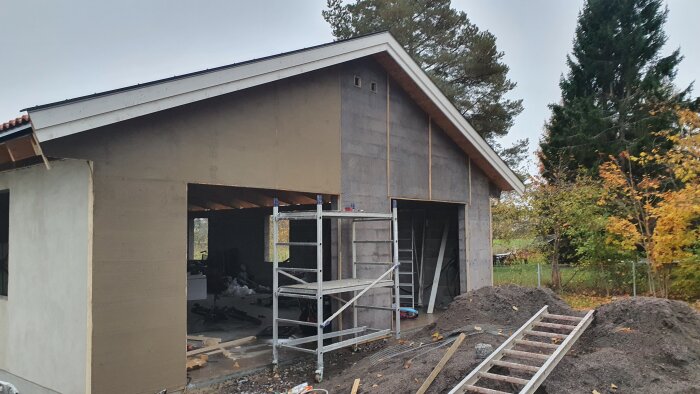 Garage under konstruktion med nyputsad vägg och tak, byggställning framför öppning och stege liggande på marken, omgiven av höstträd.