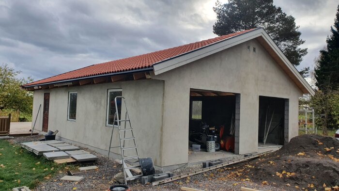 Nybyggt garage med rött tak och putsad fasad, byggmaterial och stege i förgrunden, molnig himmel i bakgrunden.