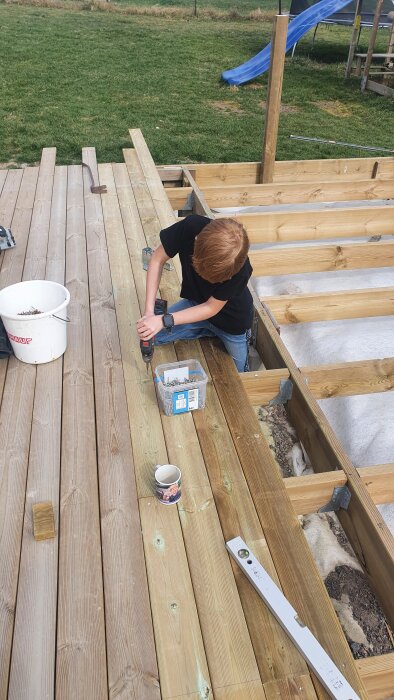 En person arbetar på en träaltan, använder en skruvdragare och har en hink med skruvar bredvid sig. Gräs och en blå rutschkana syns i bakgrunden.