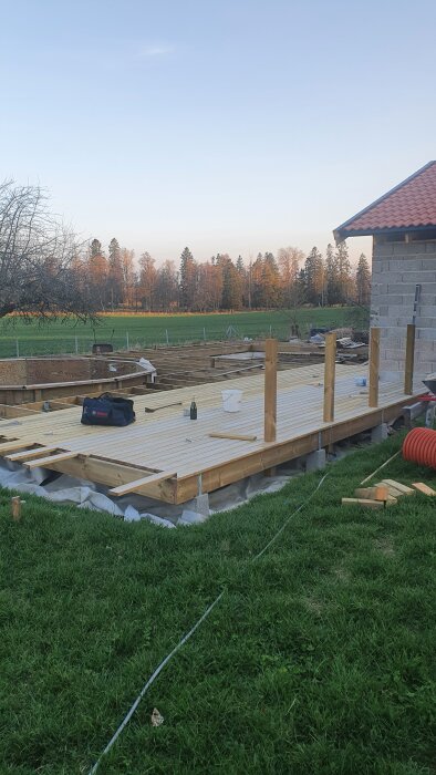 Altanbygge med använd återbrukat virke och poolkonstruktion i trä, omgiven av gräsmatta och skog i bakgrunden.