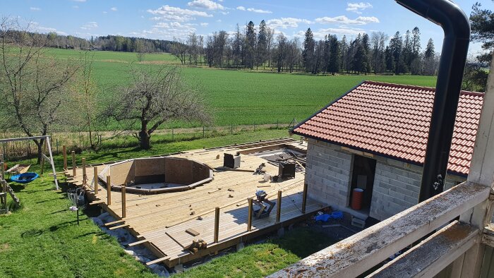 Altan med rund pool under byggnation, omgiven av grönska och ett halvtak med röda tegelpannor. Verktyg och material ligger utspridda.