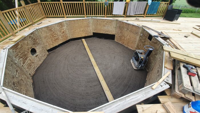 Renoverad folkpool med plywoodväggar och sandbotten, omgiven av trädäck. Träreglar och verktyg synliga på däcket.