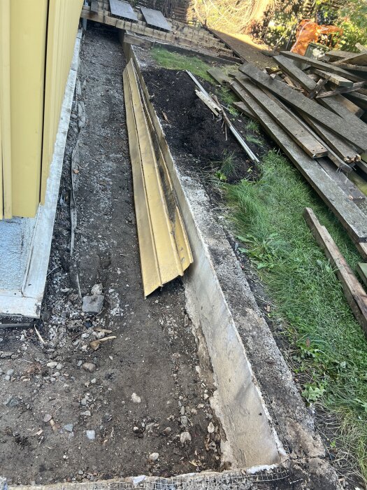 Bild av en dräneringsmur längs ett gult hus, med jordbruksmark och lösa plankor intill, i ett påbörjat trädgårdsprojekt.