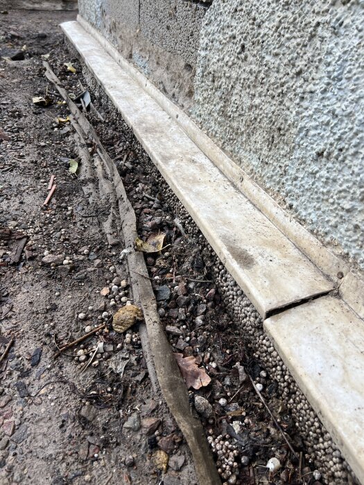 Mark med grus och rör längs husvägg, pusslande kant av stenplattor, skadad markduk, möjligt dräneringsskydd synligt i bilden.