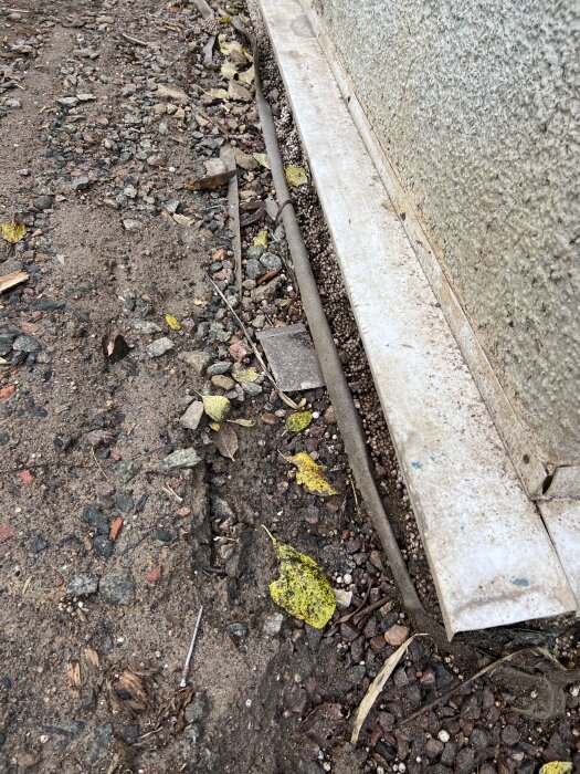 Markyta vid husvägg med sten och jord, synlig mur och avlägsnad markduk, löv och en skruv på marken.