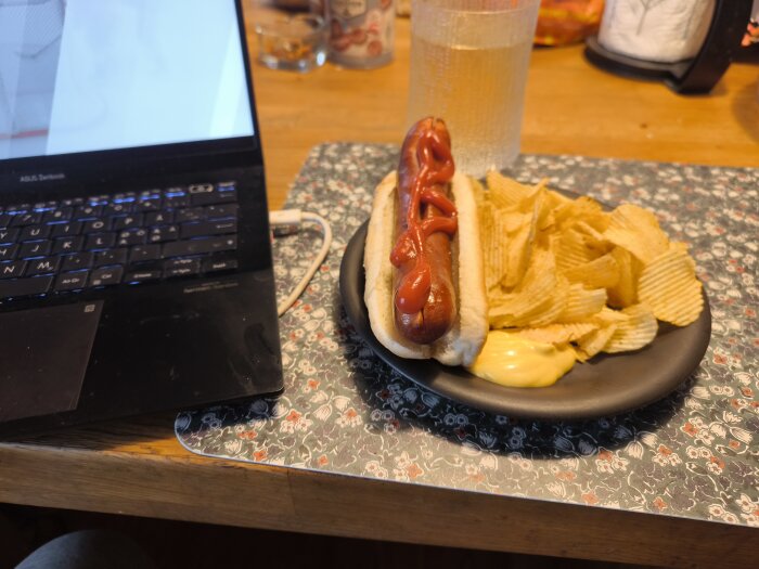 Korv i bröd med ketchup, chips och dipp på en tallrik bredvid en bärbar dator på ett bord.