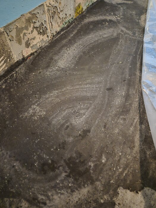 Sliten betongyta med märken efter vattning och hårda skor, omgiven av avskavd vägg och täckt med plast på ena sidan.