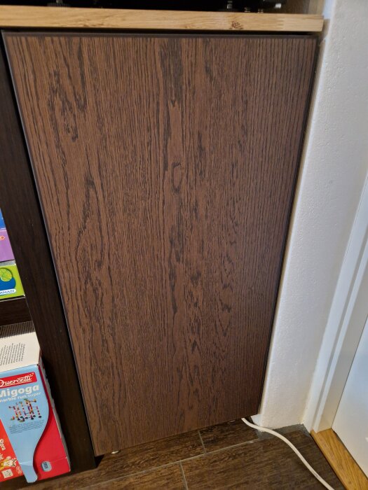 Brun träskiva monterad vertikalt ovanpå ett skåp, omgiven av vit vägg och annan möbel, syns delvis trägolv och en vit kabel nedanför.