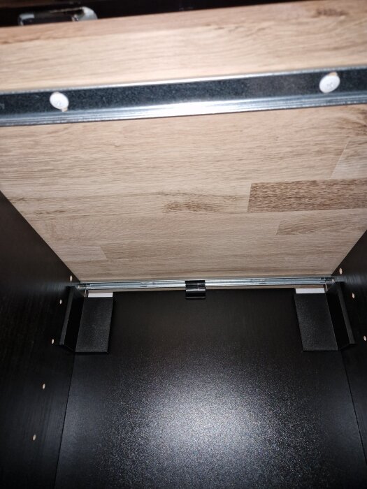 Undersida av stavlimmad ekplanka monterad med metallbeslag i ett möbel eller skåp, visar ådring och färgmatchning med annan inredning.