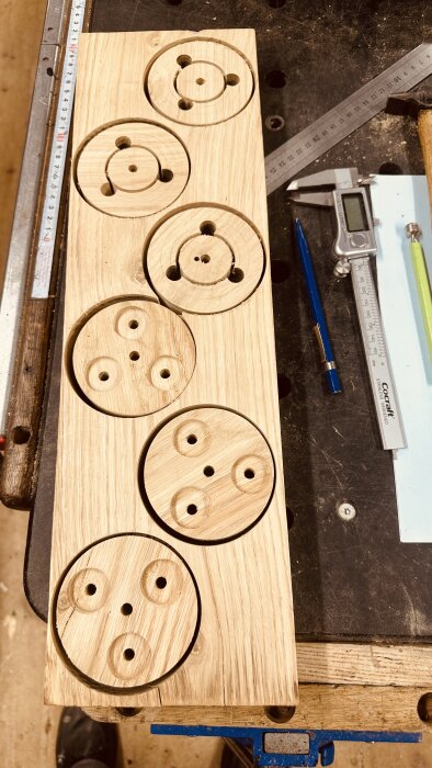 Träbitar i ek med utskurna cirkulära adaptrar och hål, inlagda i arbetsbänk, omgivna av verktyg och linjaler.