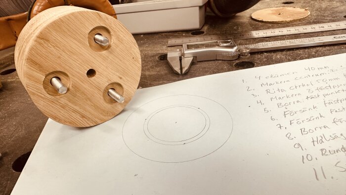 Ekadapter med tre skruvar och centrumhål, placerad på arbetsbänk med ritning och skjutmått bredvid.