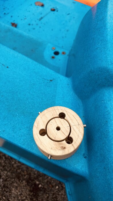 Träadapter med borrhål och skruvar placerad mot en ljusblå bakgrund, del av en lösning för att ansluta bågens rör till båtens skruvfästen.