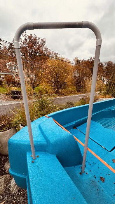 Blå trampbåt med grått rostfritt rörformat altanräcke monterat på båtens akter. Bakgrund med höstträd och grusväg.