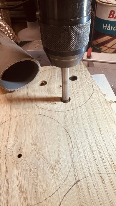 Bänkgrillen Bosch PBD-40 borrar noggrant genom en ekbräda med flera markerade cirklar för hål, med ett plaströr och en burk i bakgrunden.