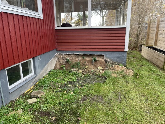 Husvägg och uterum med synlig grund där rabatten har avlägsnats, marken delvis uppgrävd och blottlagd jord syns.