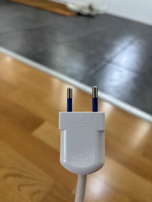 Vit strömkontakt med CE-märke mot en bakgrund av trä- och klinkergolv.