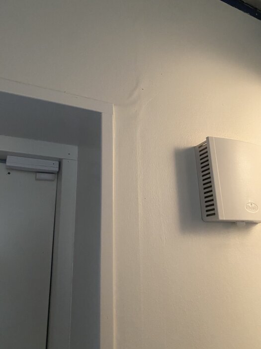 Vägg vid dörr med ojämn yta, bredvid en vit ventil, i behov av renovering eller reparation.