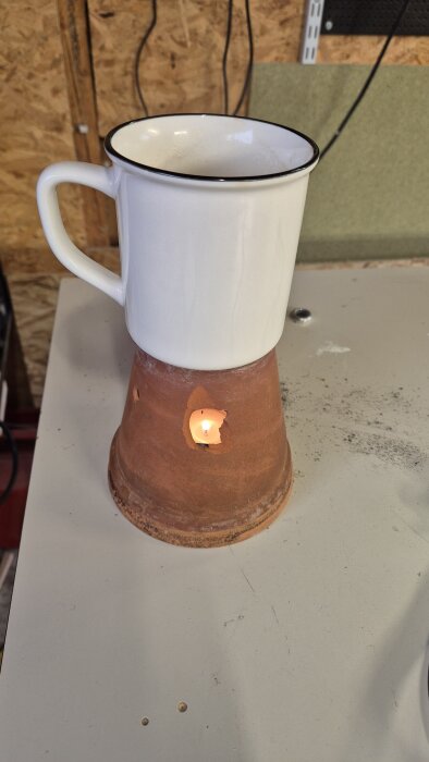 Vit kaffekopp på hemmagjord värmare av uppochnedvänd lerkruka med ljus i, placerad på bord i garage.