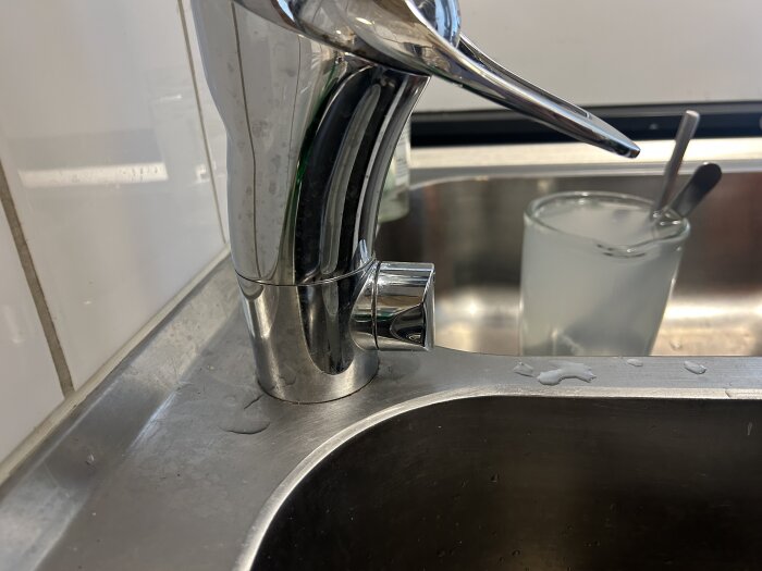 Köksblandare över en rostfri diskbänk med ett glas med vatten och två skedar i bakgrunden.