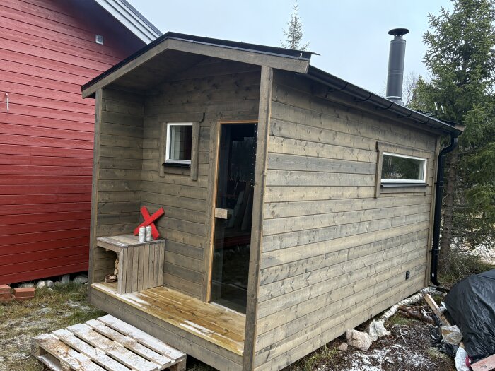 En liten bastubyggnad i trä med en svart skorsten, belägen bredvid en röd byggnad och nära ett trädsnår.