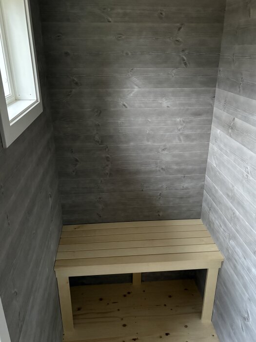Träpanelklädd bastuvägg med enkel träbänk i hörnet och ett fönster ovanför.
