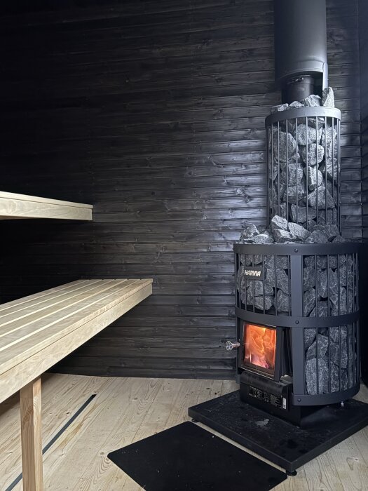 Vedeldad bastu med Harvia-ugn, omgiven av bastustenar. Vänster sida visar en träbänk, mörka väggar och trägolv.