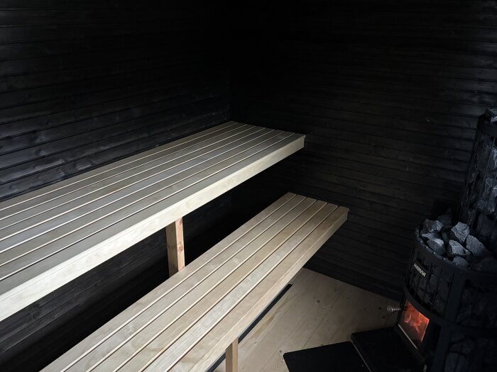 Inuti en bastu med två träbänkar och en vedeldad bastuugn fylld med stenar. Svarta väggar skapar en mysig atmosfär.