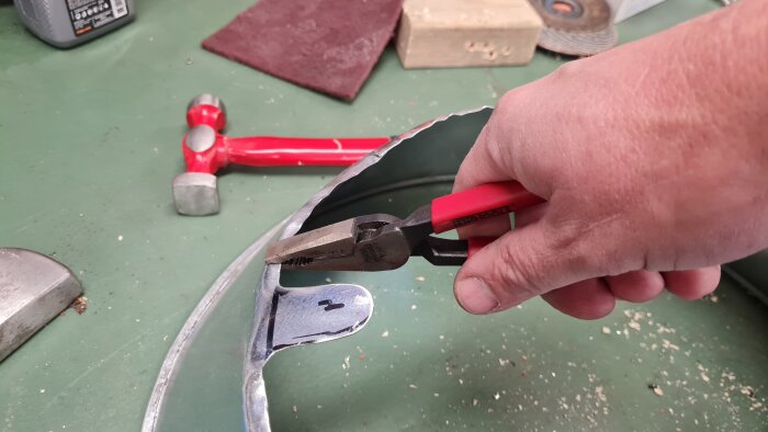 Hand arbetar med plåtsax på en metallkant, verktyg och material synliga i bakgrunden på en arbetsbänk.