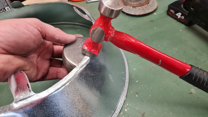 Hand håller en metalldel mot en ventilationsreducering medan en röd hammare används för formning.