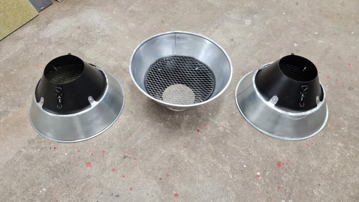 Tre plåttrattar på ett golv, två med svarta toppar och en helt i metall. Används för svetsutsug, nyligen formade och klara för test.