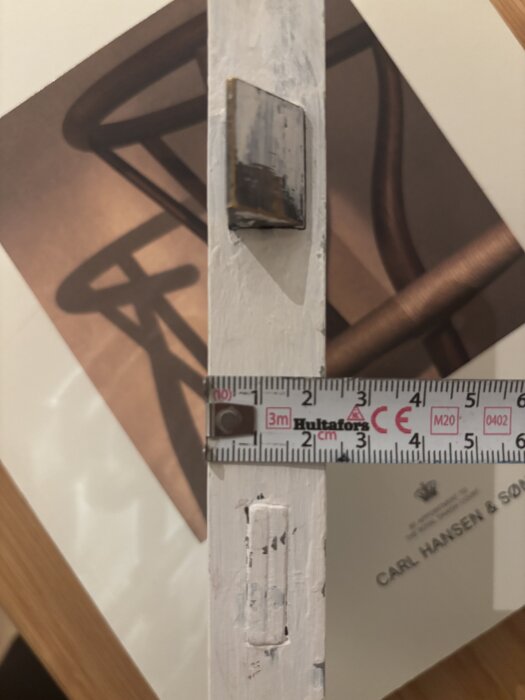 En närbild av en dörrkant med en mätstock som visar bredden, samt en metallplatta och en utskuren del av dörren.